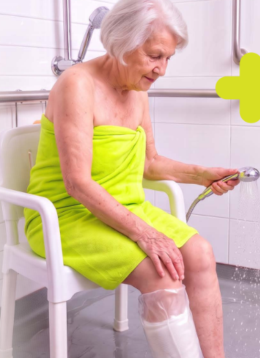 Older woman seated on a shower chair wearing a VitaPlus leg cast protector while showering.